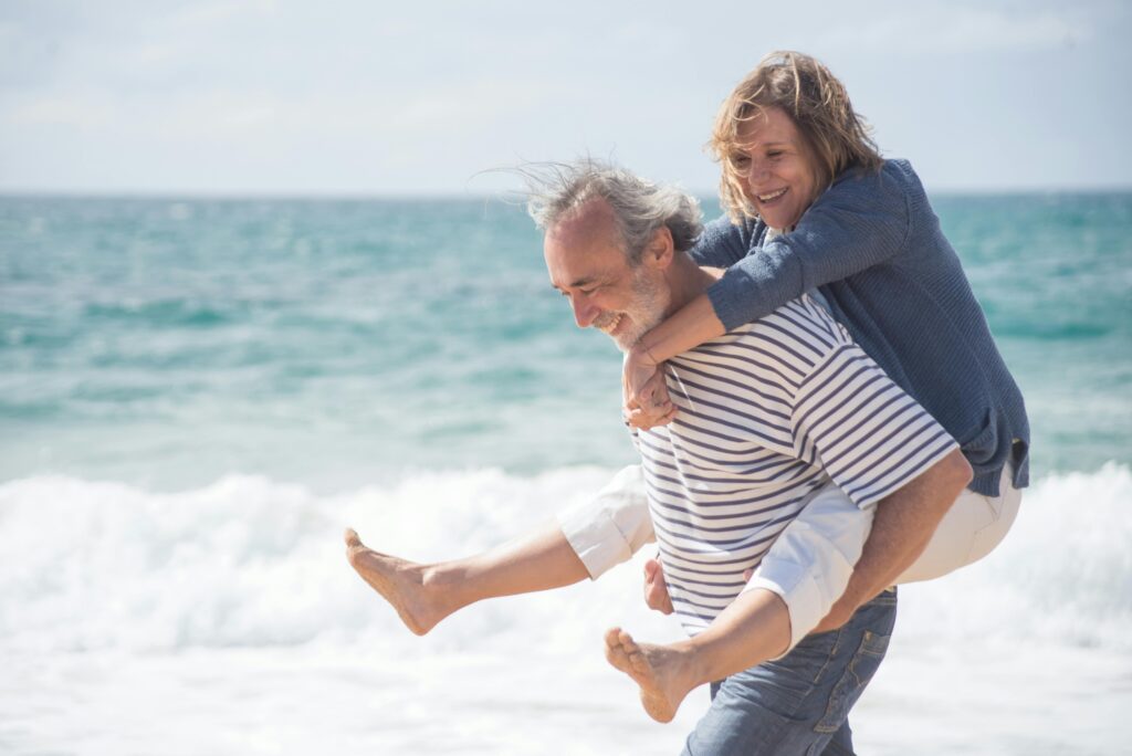 Couple senior souriant enlacé sur une plage, face à la mer, symbolisant complicité et bonheur.